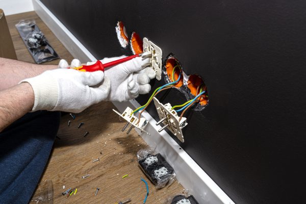 An electrician uses a red screwdriver to screw a double grounded electrical socket in a row to the electrical box in the black wall of the room.
