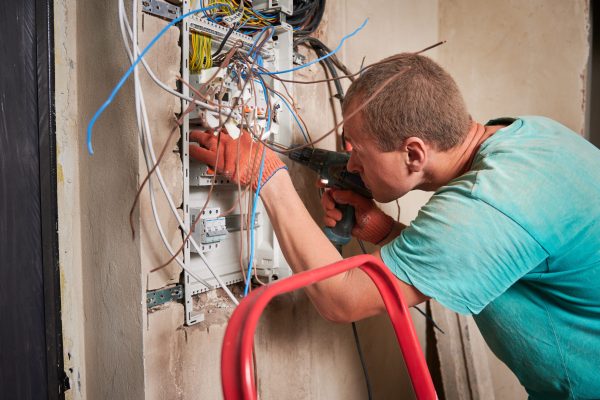 Side view of man in work gloves fixing electrical switchboard with electric drill in apartment under renovation. Electrician repairing household electrical panel with wires and switches.